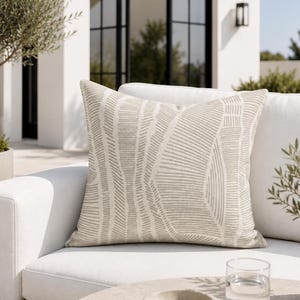 May include: A square throw pillow featuring a light beige fabric base with a repeating pattern of dark beige, thin, vertical, and curved lines. The pillow has a structured, plump appearance and is positioned on a white outdoor sofa cushion.