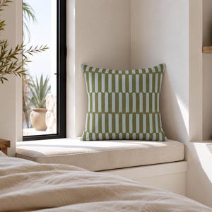May include: A square throw pillow featuring a repeating geometric pattern of vertical olive green and light blue bars. The design consists of alternating colored columns with rounded ends, creating a structured, symmetrical visual effect.