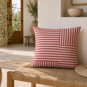 May include: A square throw pillow featuring a geometric pattern of alternating red and white stripes. The design is divided into four quadrants, with the stripes oriented horizontally in two sections and vertically in the other two sections.