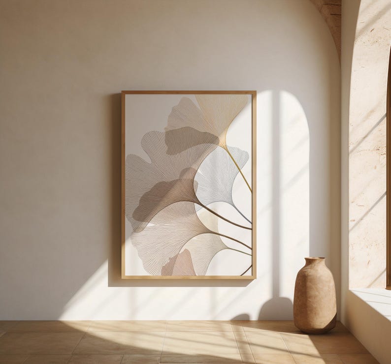 May include: A framed art print featuring a botanical design of stylized ginkgo leaves in shades of beige, brown, and white. The artwork is displayed on a white wall, with a wooden frame. A decorative vase sits on the floor.