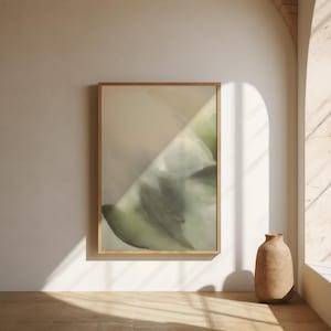 May include: A framed abstract art print in a neutral colour scheme. The artwork features blended shades of green and beige, creating a calming and organic aesthetic. A decorative, light brown vase sits on the floor.