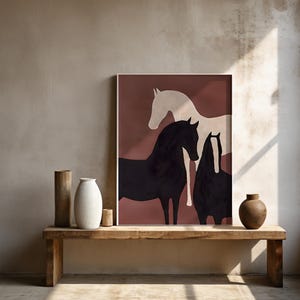 K&ouml;nnte beinhalten: Gerahmter Kunstdruck mit drei stilisierten Pferden in Schwarz und Wei&szlig; vor einem ged&auml;mpften rotbraunen Hintergrund. Das Kunstwerk wird auf einer rustikalen Holzbank zusammen mit dekorativen Vasen und einem kleinen Holzbecher pr&auml;sentiert.