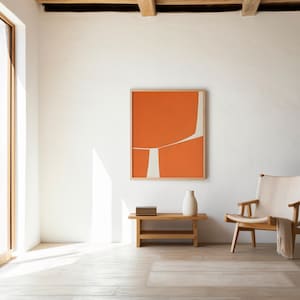 May include: A light and spacious room featuring an orange and white abstract artwork in a wooden frame. A wooden bench holds a white vase and books. A wooden chair with a cream seat and a blanket is in the foreground. The room has a wooden floor.
