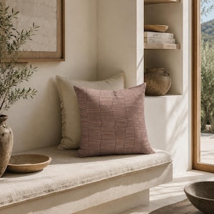 May include: A square throw pillow in a muted mauve color featuring a pattern of thin, dark, wavy horizontal lines. The pillow is positioned in front of a plain, light beige square pillow, both resting on a flat, light-colored bench surface.