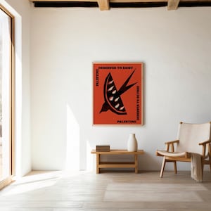May include: A framed poster with a red background and a black bird graphic. The poster includes the text "Palestine Deserves to Exist" and "Palestine Deserves to Be Free". The poster is displayed in a bright, minimalist room with a wooden chair and table.