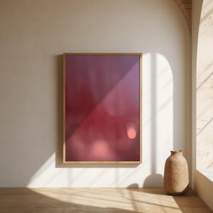 Op de afbeelding: Een ingelijste abstracte kunstprint met een wazig, gradiëntontwerp in rood- en paarstinten. Het kunstwerk wordt tentoongesteld op een witte muur, met een neutrale vaas en natuurlijk licht van een raam. De houten lijst voegt warmte toe.