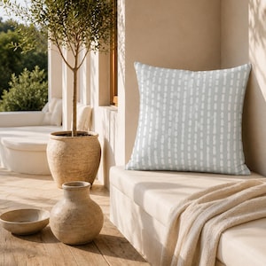 May include: A square throw pillow featuring a light sage green background with a repeating pattern of vertical, dashed white lines. The pillow is positioned on a light-colored bench, showcasing a soft fabric texture and a clean, geometric design.