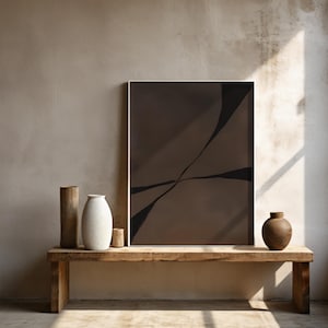 May include: A framed abstract art print with a brown and black design, displayed on a wooden bench. Accompanying the artwork are three vases and a small wooden cup. The room has a neutral color palette with natural light.