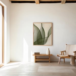 May include: A square wall art piece featuring a series of overlapping, dark green, curved line patterns that create a leaf-like design on a light cream background, enclosed within a simple, light-toned wooden frame hanging on a white wall.