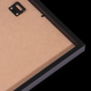 May include: A black picture frame with a brown backing. The frame has a simple, modern design. A metal sawtooth hanger is attached to the back for wall mounting. The frame is set against a black background.