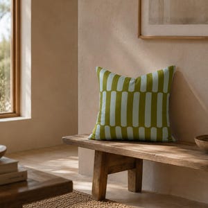 May include: A decorative square pillow with a green background and vertical light blue stripes. The pillow is placed on a rustic wooden bench. The room has a neutral color palette with natural light.