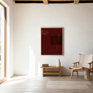 Puede incluir: Escena interior minimalista con una obra de arte abstracta enmarcada en tonos burdeos y marrón. Un banco de madera sostiene un jarrón blanco y libros. Una silla de color claro con una manta drapeada está cerca. La habitación tiene una ventana grande.