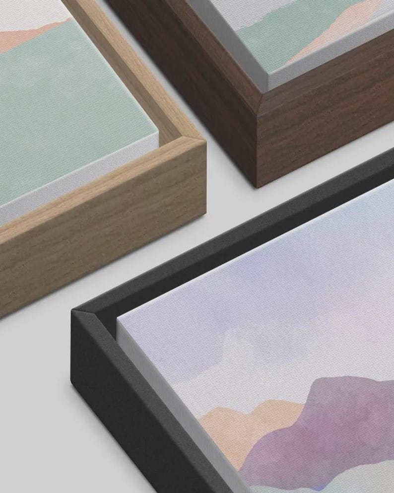 May include: Close-up of three framed canvas art prints. The frames are in natural wood, dark brown, and black. The artwork features a watercolor landscape in pastel colors, including lavender, peach, and mint green.