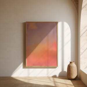 May include: A framed abstract art print with a gradient of colors, including shades of purple, orange, and pink. The artwork is displayed on a white wall, with a neutral-toned vase on the floor. Natural light casts shadows across the scene.