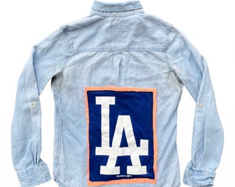 May include: Light blue denim shirt with long sleeves. The back features a large blue rectangle with the white letters "LA" and an orange border. The shirt has buttoned cuffs and a classic collar.
