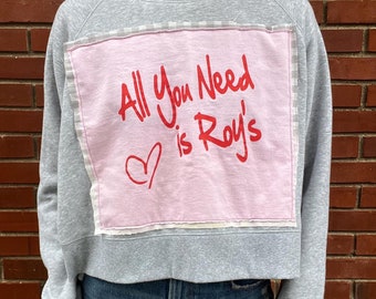 May include: A gray cropped sweatshirt with a pink patch that says "All You Need is Roy's" in red lettering with a heart.