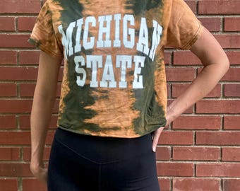 May include: A tie-dye crop top in shades of brown and green with the words "MICHIGAN STATE" in white block letters. The top is paired with black athletic shorts. The background is a red brick wall.