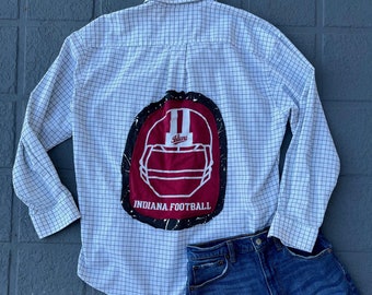 May include: A white button-down shirt with a black and white checkered pattern. The back of the shirt has a red and white patch with a football helmet and the text "Indiana Football". The shirt is paired with blue denim shorts.
