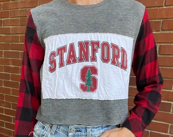 May include: A cropped grey and red long-sleeved shirt with a white patch that says "STANFORD" in red with a red "S" and a green pine tree inside. The sleeves are a red and black buffalo plaid.