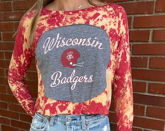 May include: A cropped long-sleeve shirt with a red and yellow tie-dye pattern. The shirt features a gray patch with the words "Wisconsin Badgers" and a helmet graphic. The shirt is paired with light blue denim jeans.