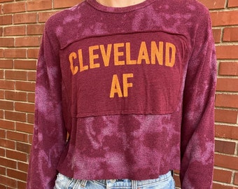 May include: Burgundy long-sleeve crop top with tie-dye effect. The shirt features the words "CLEVELAND AF" in orange block letters across the chest. The top is paired with light blue denim jeans.