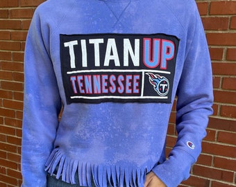 May include: A purple cropped sweatshirt with a frayed hem. The sweatshirt has a black patch with the words "TITAN UP" and "TENNESSEE" in white and red lettering. The patch also features a blue and red logo with a flame and a "T".