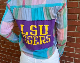 May include: A plaid button-up shirt with a purple patch on the back that says "LSU TIGERS" in yellow letters. The shirt is worn over white jeans.