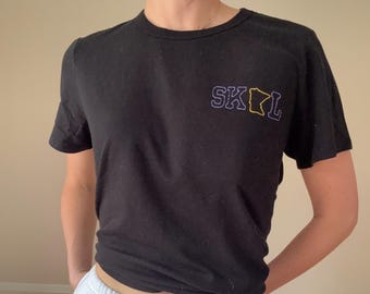 May include: A black t-shirt with a purple and yellow embroidered design that says "SKOL" with an outline of the state of Minnesota.