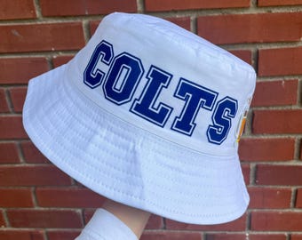 May include: White bucket hat with the word "COLTS" in blue block letters across the front. The hat has a wide brim and is made of a textured fabric. The background is a red brick wall.