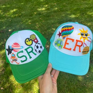 Op de afbeelding: Twee kleurrijke trucker caps met patches. De groene cap heeft een witte kroon met patches van een planeet, een ster, een astronaut, een voetbal, een Pac-Man, frietjes en een pizzapunt. De blauwe cap heeft een witte kroon met patches van een luchtballon, een auto, een zon met een zonnebril, een honkbal en een hamburger. De tekst "SR" staat op de groene cap en "ER" op de blauwe cap.