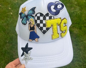 May include: White mesh trucker hat with various colorful patches, including a butterfly, a checkered heart, a record, a singer, and the initials "TS" in yellow with gold glitter.