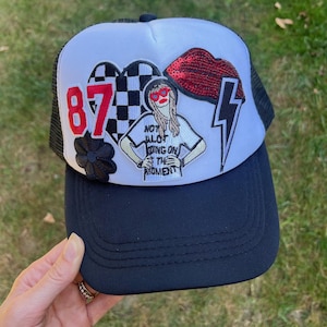 Puede incluir: Gorra de camionero blanca con una malla negra en la parte posterior. La gorra tiene varios parches, incluyendo un corazón rojo con un patrón de cuadros blanco y negro, un labio rojo de lentejuelas, un rayo y una flor negra. La gorra también tiene un parche blanco con una mujer que lleva gafas de sol y una camiseta blanca con el texto "Not Alot Going On The Moment". La gorra tiene el número "87" en rojo en la parte delantera.