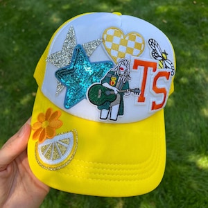 Peut inclure: Casquette de camionneur jaune avec un filet blanc à l'arrière et divers patchs colorés, dont une étoile, un cœur, une abeille, une figurine jouant de la guitare et les lettres "TS".