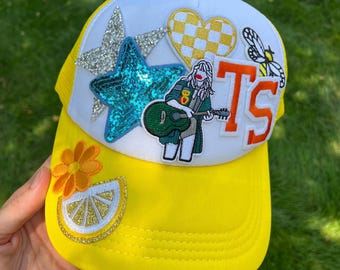 May include: Yellow trucker hat with a white mesh back and various colorful patches, including a star, a heart, a bee, a figure playing a guitar, and the letters "TS".