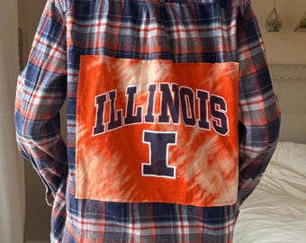 May include: Plaid flannel shirt in navy blue, red, and orange. The back features a large orange patch with the word "ILLINOIS" and the letter "I" in white block letters, with a tie-dye effect.