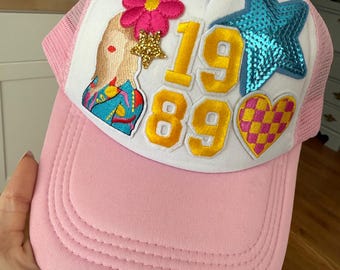 May include: Pink trucker hat with a white front panel featuring a pink flower, gold star, blue star, and a checkered heart. The hat has the numbers "1989" in gold. The hat has a pink mesh back and a pink brim.