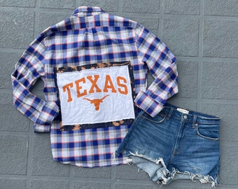 May include: A blue and white plaid flannel shirt with a white patch sewn on the back that says "TEXAS" in orange letters with a burnt orange Longhorn steer logo. The shirt is paired with a pair of light blue denim cut-off shorts.