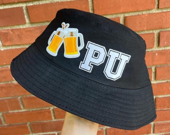 May include: Black bucket hat with the letters "PU" in white block letters. The hat also features a patch with two beer mugs. The hat is held up against a brick wall.