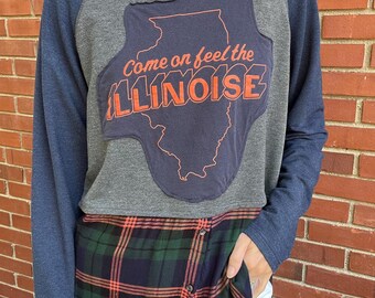 May include: A gray and navy blue long-sleeved shirt with a patchwork design. The shirt features a graphic of the state of Illinois with the text "Come on feel the ILLINOISE". The shirt is layered over a plaid shirt.