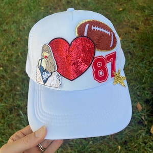 Puede incluir: Gorra de camionero blanca con un corazón rojo, una pelota de fútbol marrón y el número 87 en rojo. La gorra también tiene una estrella dorada y una imagen de dibujos animados de una mujer.