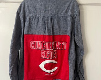 May include: A blue denim long-sleeve button-up shirt with a red patch on the back. The patch features the text "Cincinnati Reds" and a white "C" logo.