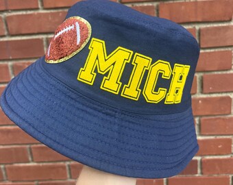 May include: A navy blue bucket hat with a yellow and gold embroidered football and the word "MICH" on the front.