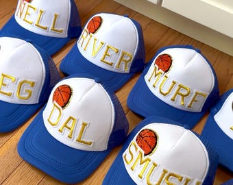 May include: A collection of blue and white trucker hats with gold embroidered names and a basketball graphic. The hats have a blue brim and mesh back, with names like "NELL", "RIVER", and "DAL" on the front.
