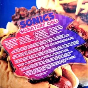 May include: A recipe card for Sonic's Signature Chili, with ingredients and instructions. The card is arrow-shaped, with a purple and red design. The chili recipe includes ground beef, spices, and vegetables. Measurements are in ounces and pounds.