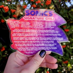 May include: A clear acrylic recipe card for Sonic's Signature Chili, held by a hand. The card features a purple and pink design with the recipe's ingredients and instructions. The background is blurred with red flowers.