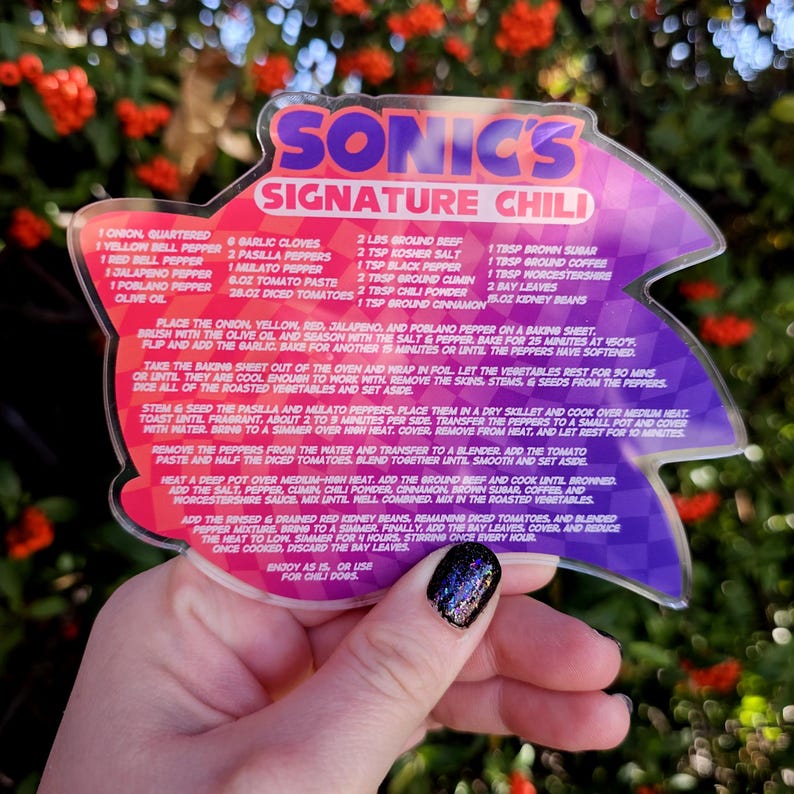 May include: A clear acrylic recipe card for Sonic's Signature Chili, held by a hand. The card features a purple and pink design with the recipe's ingredients and instructions. The background is blurred with red flowers.