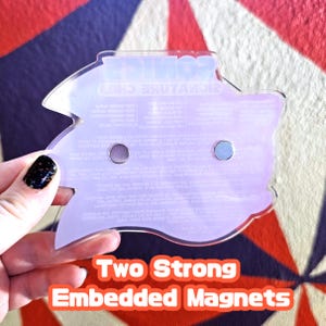 May include: A clear, Sonic the Hedgehog-shaped magnet with embedded magnets. The magnet has text and is held against a colourful background. The text "Two Strong Embedded Magnets" is at the bottom of the image.