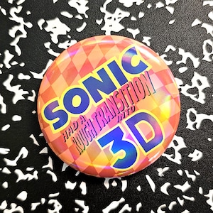 Könnte beinhalten: Ein runder Button mit einem karierten orange-rosa Hintergrund. Der Button trägt den Text "SONIC HAD A ROUGH TRANSITION INTO 3D" in blauer und rosa Schrift. Holografischer Effekt.