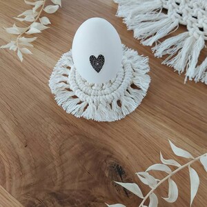 Op de afbeelding: Wit ei met een zwart hartontwerp op een witte gehaakte eierhouder met franjes. De eierhouder staat op een houten oppervlak.