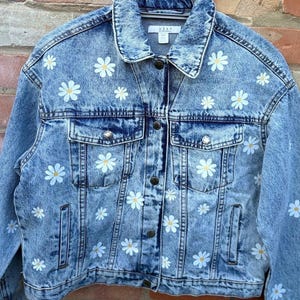 Puede incluir: Una chaqueta de mezclilla azul con lavado ácido que presenta un patrón repetitivo de margaritas blancas con centros amarillos. La chaqueta tiene cuello clásico, mangas largas, dos bolsillos en el pecho con botones metálicos y cierre frontal con la etiqueta WEAR.
